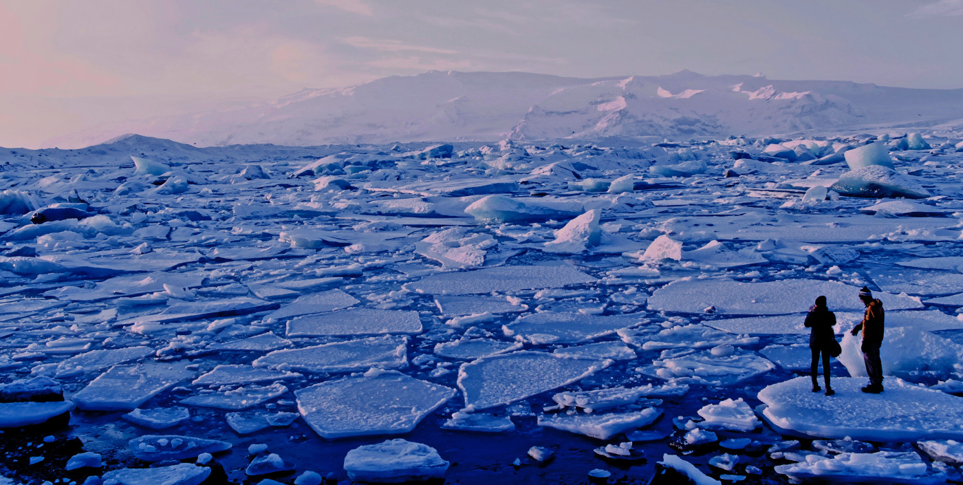 Two people stand on a small sheet of ice surrounded by a many other small sheets of ice on a body of water.