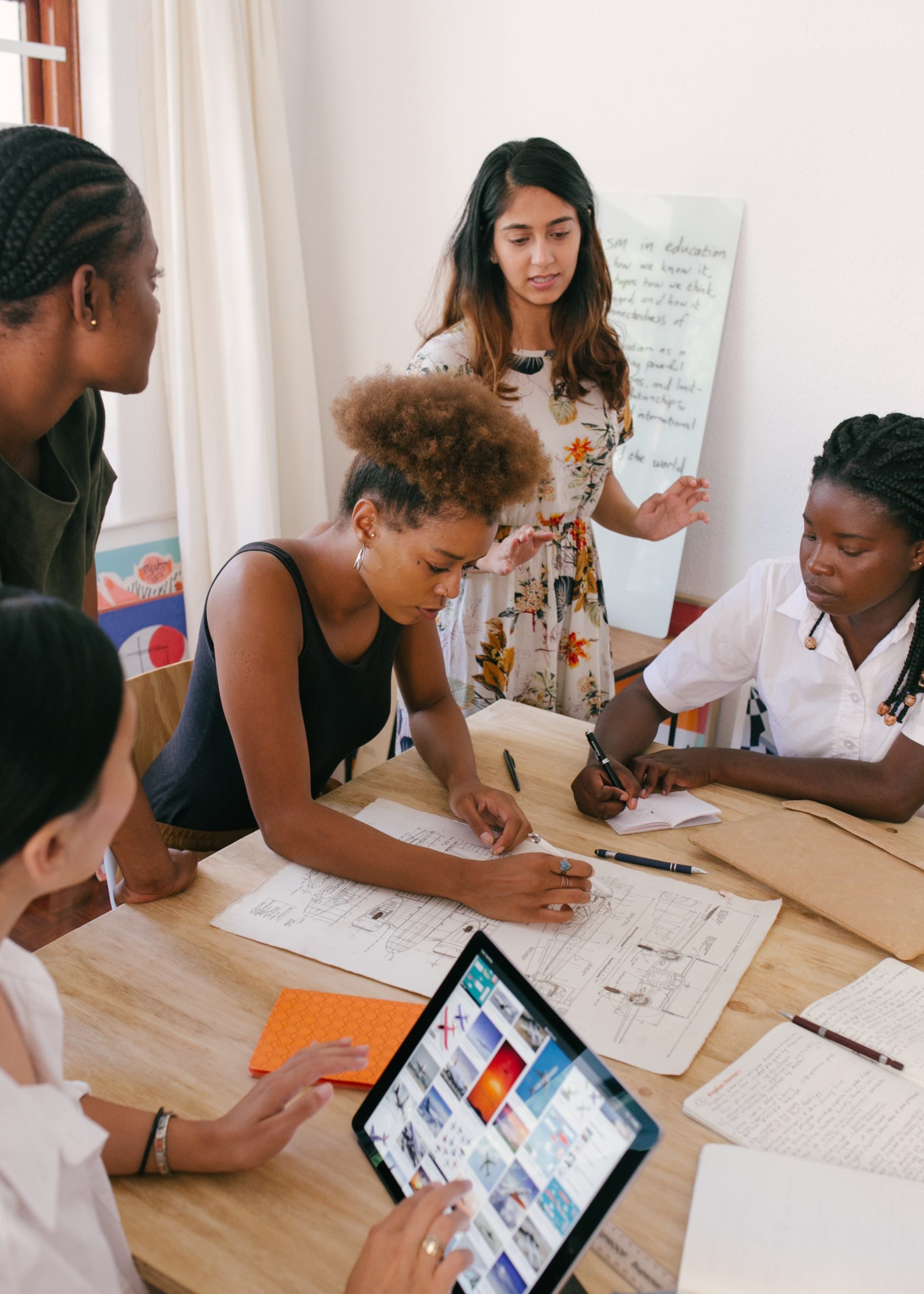 Three people sit and two people stand around a table. One person is speaking. One seated person works on a laptop, one takes notes, one works on a design plan.