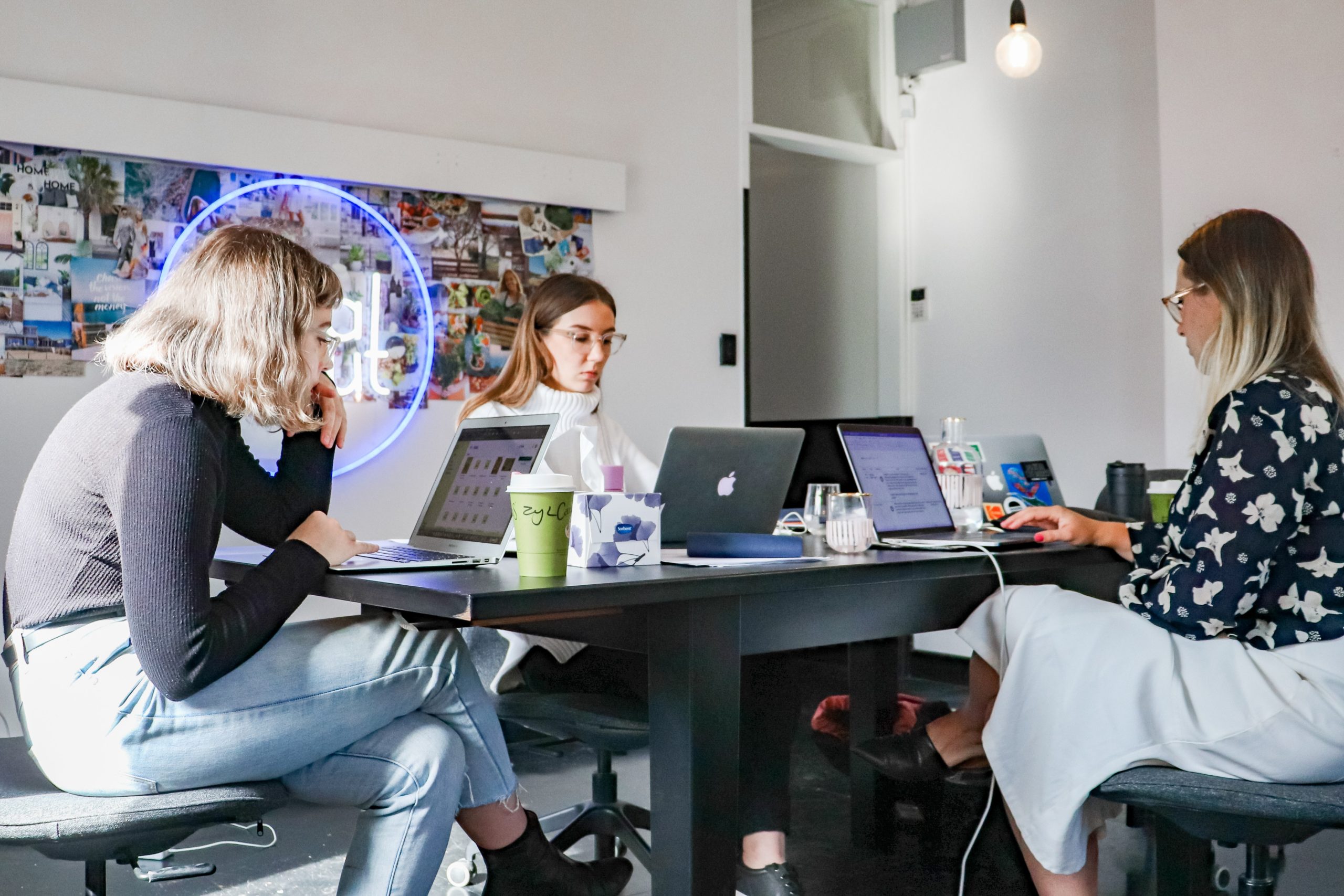 Three women seated at a table work at open laptops.