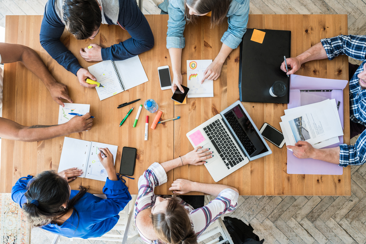 A photograph from above of six people sitting around a desk, taking notes, and working on a laptop.