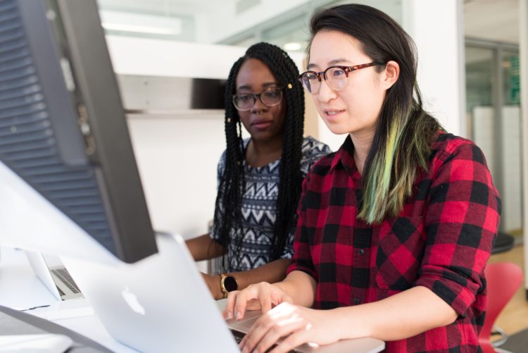 Two women with open laptops in front of them. They both look at the screen of one laptop.