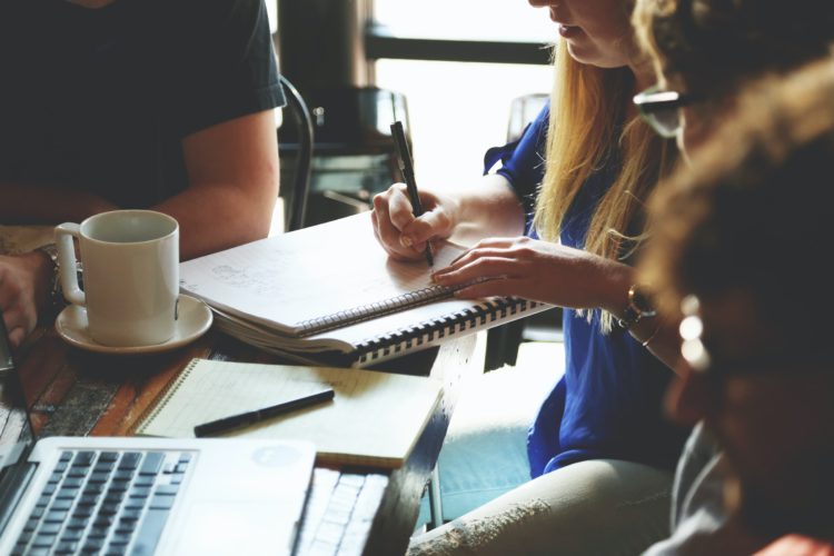 Four people seated around a table. One person writes in a notebook. An open laptop is in front of another person.