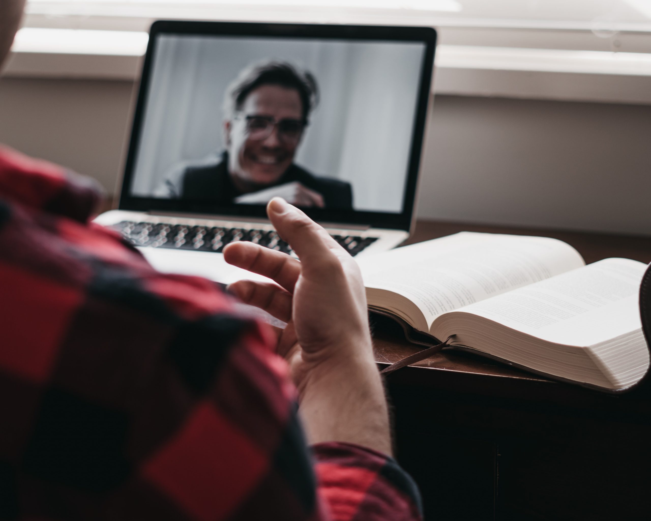 A man sits in a chair at a desk. An open laptop in front of him displays another man on video. An open book is next to the laptop.