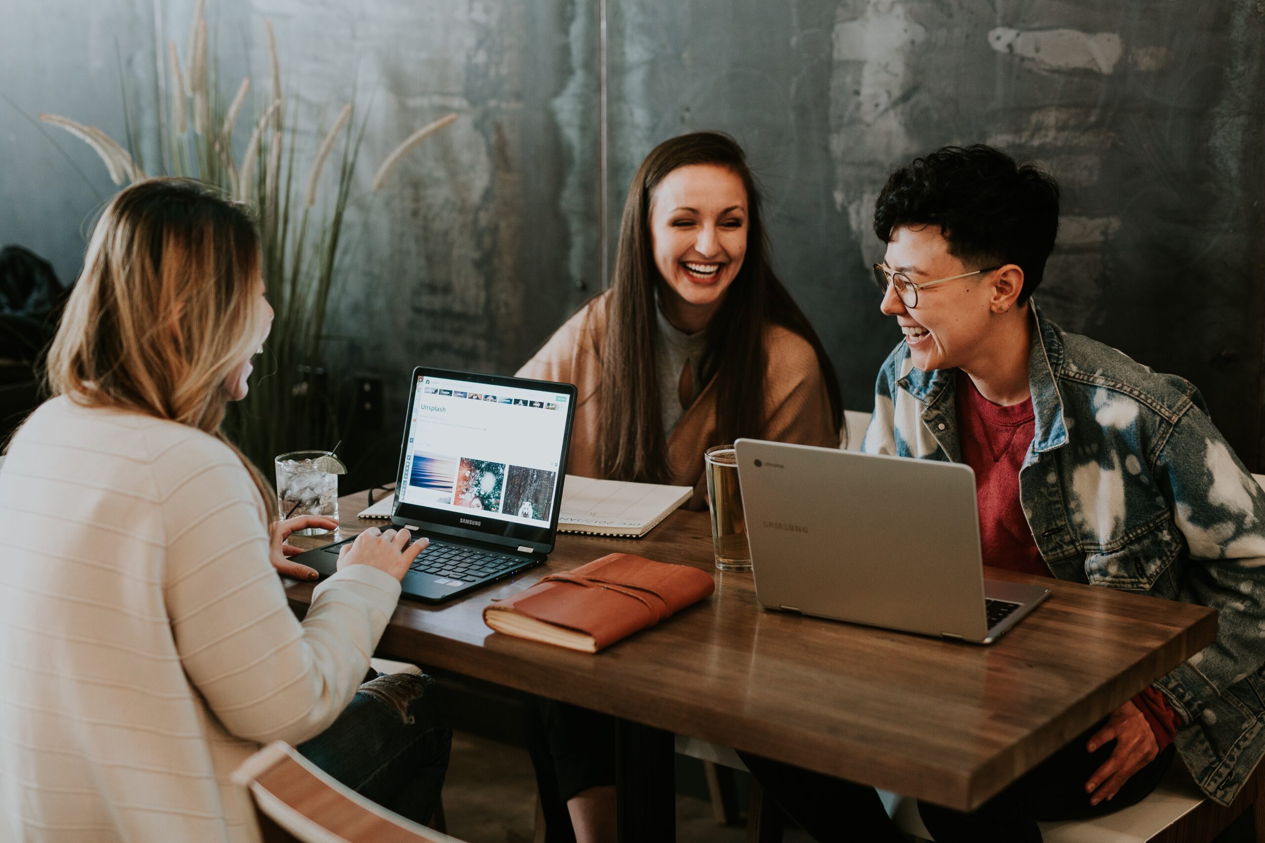Three smiling people sit at a table. Two of the people have an open laptop in front of them. The third person has a notepad in front of her.